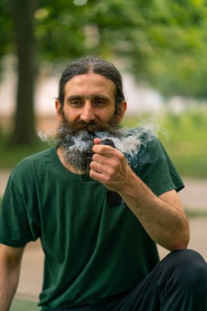 Foto de Un hombre mayor con pelo largo y barba gris fuma pipa en un parque de la ciudad con árboles en el fondo - Imagen libre de derechos