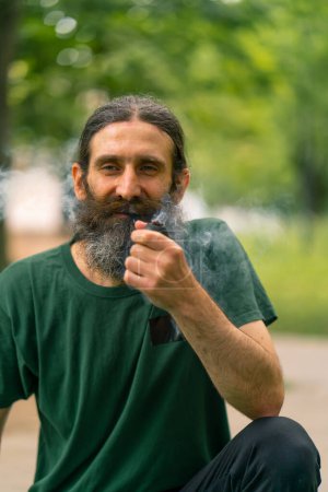 Photo for An older man with long hair and gray beard smokes pipe in a city park with trees in the background - Royalty Free Image