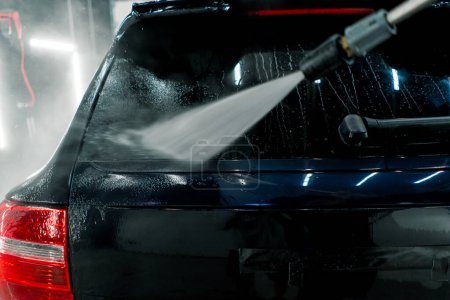 Photo for Close-up of a male car wash employee washing the rear window of a black luxury car with high-pressure washer - Royalty Free Image