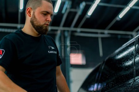 Foto de Un empleado de lavado de autos masculino aplica detergente de lavado de autos a un auto de lujo negro usando pistola en la caja de lavado de autos - Imagen libre de derechos