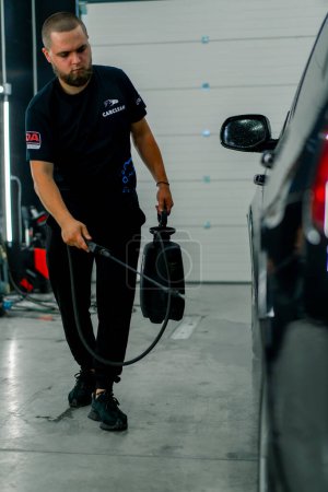 Foto de Un empleado de lavado de autos masculino aplica detergente de lavado de autos a un auto de lujo negro usando pistola en la caja de lavado de autos - Imagen libre de derechos