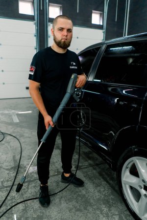 Téléchargez les photos : Portrait d'un ouvrier de lavage de voiture tenant un pistolet de lavage haute pression sur le fond de voiture noire de luxe - en image libre de droit