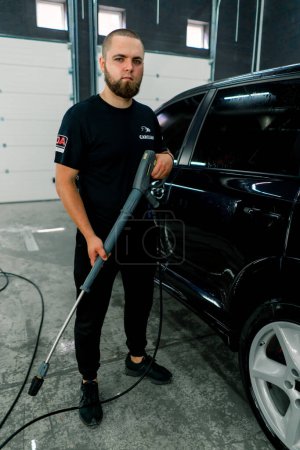 Téléchargez les photos : Portrait d'un ouvrier de lavage de voiture tenant un pistolet de lavage haute pression sur le fond de voiture noire de luxe - en image libre de droit
