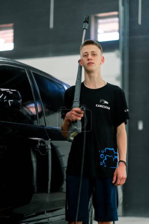 Téléchargez les photos : Portrait d'un ouvrier de lavage de voiture tenant un pistolet de lavage haute pression sur son épaule sur le fond de voiture noire de luxe - en image libre de droit