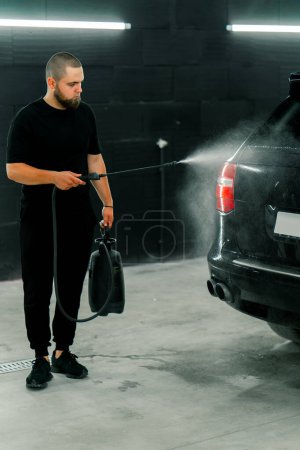 Photo for A male car wash employee applies car wash detergent to a black luxury car using spray gun in the car wash box - Royalty Free Image