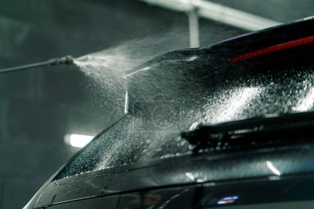 Téléchargez les photos : Gros plan de la vitre arrière d'une voiture noire au moment d'appliquer le lavage de voiture d'un vaporisateur dans une boîte de lavage de voiture - en image libre de droit