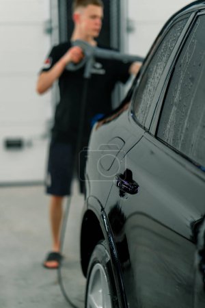 Photo for A male car wash employee washes a black luxury car with high-pressure washer in the car wash bay - Royalty Free Image