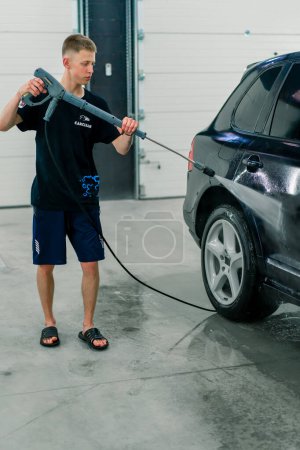 Téléchargez les photos : Un employé de lavage de voiture masculin lave une voiture de luxe noire avec laveuse haute pression dans la salle de lavage de voiture - en image libre de droit