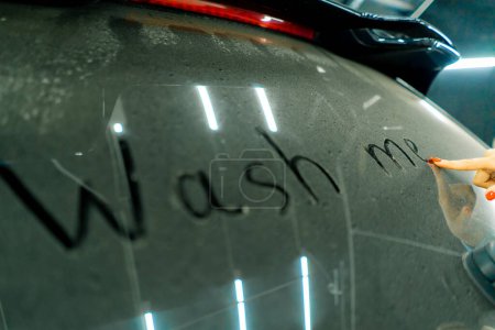 Photo for A close-up of a woman's hand with a red manicure writing "wash me" on the dirty rear window of a luxury blue car in front of car wash - Royalty Free Image