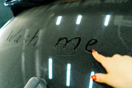 Foto de Un primer plano de la mano de una mujer con una manicura roja escrito "lávame" en la ventana trasera sucia de un coche azul de lujo en frente de lavado de coches - Imagen libre de derechos
