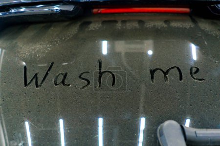 Photo for A close-up of a wash me sign on the dirty rear window of a luxury blue car in front of car wash - Royalty Free Image