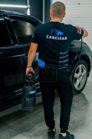 Photo for A male car wash employee applies car wash detergent to a black luxury car using spray gun in the car wash box - Royalty Free Image