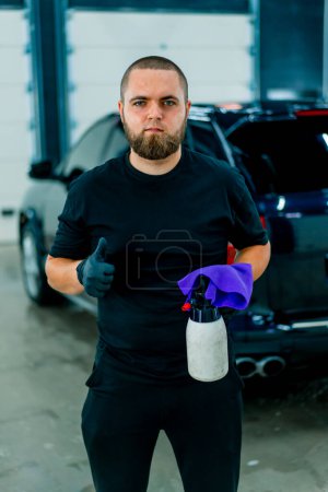 Téléchargez les photos : Portrait d'un ouvrier de lavage de voiture mâle avec un pistolet et un chiffon en microfibre dans ses mains qui montre un signal pouce en arrière-plan de voiture bleue de luxe - en image libre de droit