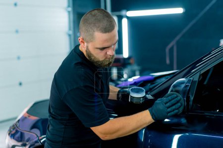 Foto de Trabajador masculino enfocado del lavado del coche en guantes negros pule el espejo de un coche azul de lujo usando el concepto del cuidado del coche de la esponja del pulido - Imagen libre de derechos