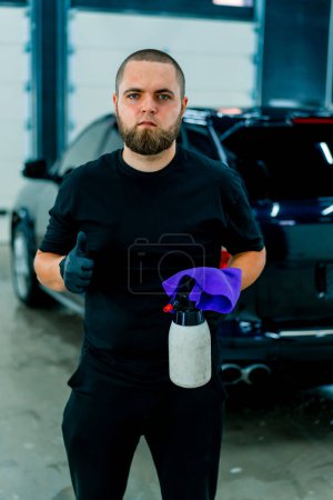 Foto de Retrato de un trabajador de lavado de coches masculino con una pistola de pulverización y un paño de microfibra en sus manos que muestra una señal de pulgar en el fondo del coche azul de lujo - Imagen libre de derechos