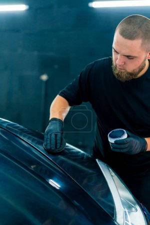 Téléchargez les photos : Un travailleur de lavage de voiture concentré dans des gants noirs polit le capot et le phare d'une voiture noire de luxe avec une éponge de polissage de concept de soins de voiture - en image libre de droit