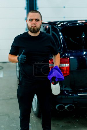Photo for Portrait of a male car wash worker with a spray gun and a microfiber cloth in his hands who shows a Thumb signal in background of luxury blue car - Royalty Free Image