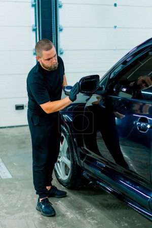 Téléchargez les photos : Focused Homme lavage de voiture travailleur dans des gants noirs polit le miroir d'une voiture bleue de luxe en utilisant le concept de soins de voiture éponge de polissage - en image libre de droit
