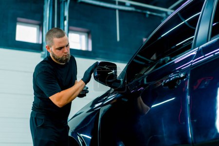 Téléchargez les photos : Focused Homme lavage de voiture travailleur dans des gants noirs polit le miroir d'une voiture bleue de luxe en utilisant le concept de soins de voiture éponge de polissage - en image libre de droit
