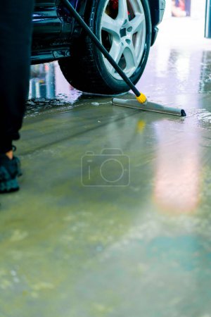 Téléchargez les photos : Gros plan d'un préposé au lavage de voiture retirant de l'eau et de la mousse du sol dans une boîte de lavage de voiture avec une serpillière après le travail de la journée - en image libre de droit