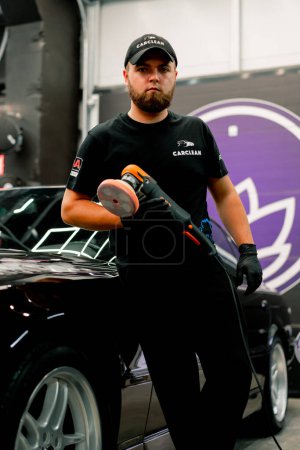 Foto de Retrato de un trabajador de lavado de coches masculino con una máquina de pulir en sus manos en el fondo de un coche de lujo negro - Imagen libre de derechos