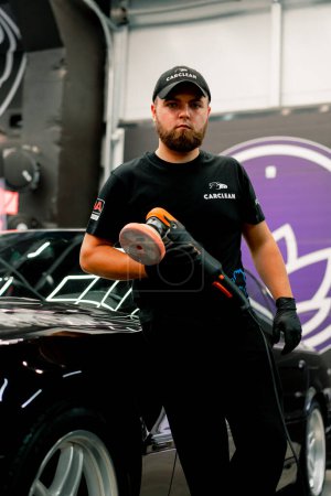 Téléchargez les photos : Portrait d'un ouvrier de lavage de voiture mâle avec une machine à polir dans ses mains sur le fond de voiture de luxe noire - en image libre de droit