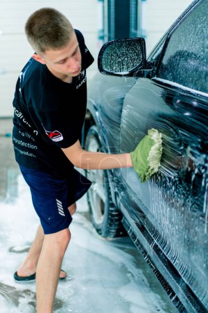 Photo for A male car wash worker uses a green washcloth to wash black luxury car with car wash shampoo - Royalty Free Image