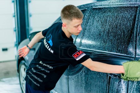 Téléchargez les photos : Un employé de lavage de voiture utilise un gant de toilette vert pour laver une voiture de luxe noire avec un shampooing de lavage de voiture - en image libre de droit