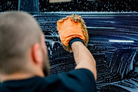 Téléchargez les photos : Gros plan d'un ouvrier de lavage de voiture utilisant un chiffon en microfibre pour laver une voiture de luxe noire avec un shampooing de lavage de voiture - en image libre de droit