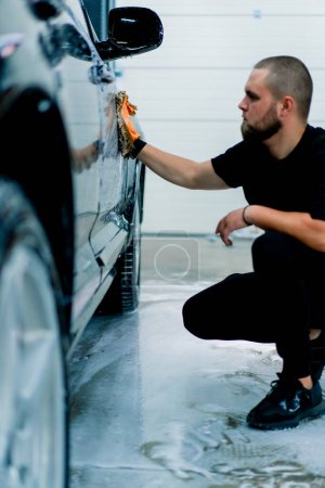 Téléchargez les photos : Un travailleur de lavage de voiture utilise un chiffon en microfibre pour laver le phare avant de la voiture de luxe noire avec shampooing de lavage de voiture - en image libre de droit