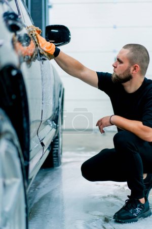 Foto de Un trabajador de lavado de coches utiliza un paño de microfibra para lavar el faro delantero del coche de lujo negro con champú de lavado de coches - Imagen libre de derechos