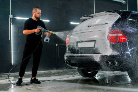 Foto de Un empleado de lavado de autos masculino aplica espuma de lavado de autos a un auto negro de lujo usando pistola en la caja de lavado de autos - Imagen libre de derechos