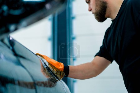 Téléchargez les photos : Un travailleur de lavage de voiture utilise un chiffon en microfibre pour laver le phare avant de la voiture de luxe noire avec shampooing de lavage de voiture - en image libre de droit
