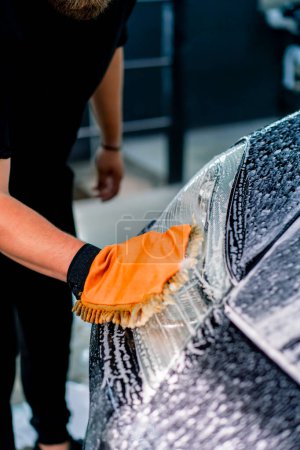 Foto de Primer plano de un trabajador de lavado de coches utilizando un trapo de microfibra para lavar el faro delantero de un coche de lujo negro con champú de lavado de coches - Imagen libre de derechos
