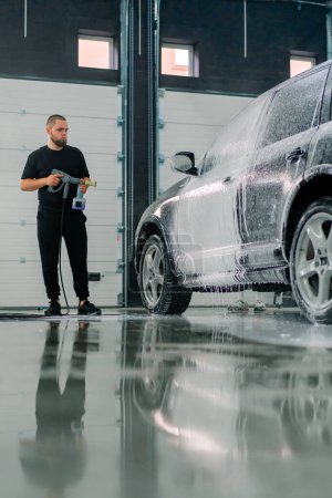 Photo for A male car wash employee applies car wash foam to a luxury black car using spray gun in the car wash box - Royalty Free Image