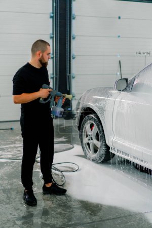 Foto de Un empleado de lavado de autos masculino aplica espuma de lavado de autos a un auto negro de lujo usando pistola en la caja de lavado de autos - Imagen libre de derechos
