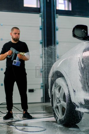 Téléchargez les photos : Un employé de lavage de voiture masculin applique de la mousse de lavage de voiture à une voiture noire de luxe à l'aide d'un pistolet dans la boîte de lavage de voiture - en image libre de droit