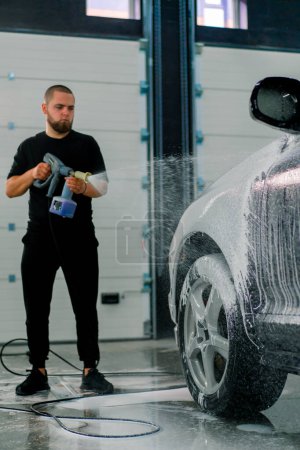 Téléchargez les photos : Un employé de lavage de voiture masculin applique de la mousse de lavage de voiture à une voiture noire de luxe à l'aide d'un pistolet dans la boîte de lavage de voiture - en image libre de droit