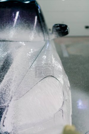 Téléchargez les photos : Gros plan de mousse de voiture sur le phare avant et le pare-chocs d'une voiture de luxe noire dans une boîte de lavage de voiture - en image libre de droit