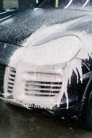 Foto de Primer plano de la espuma del coche en el faro delantero y parachoques de un coche de lujo negro en la caja de lavado de coches - Imagen libre de derechos