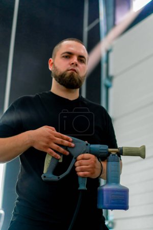 Foto de Retrato de un hombre serio con una barba de un trabajador de lavado de coches que sostiene la boquilla del generador de espuma en sus manos concepto de cuidado del coche - Imagen libre de derechos