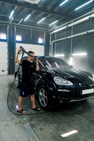 Téléchargez les photos : Un employé de lavage de voiture masculin lave une voiture de luxe noire avec laveuse haute pression dans la salle de lavage de voiture - en image libre de droit