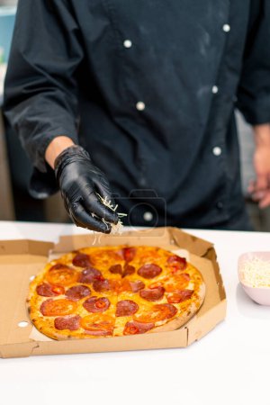 Foto de Un chef de pizza macho rocía queso mozzarella en una pizza que se encuentra en una caja de entrega en la cocina de la pizzería - Imagen libre de derechos