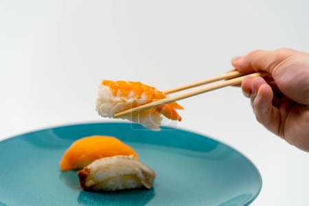 Foto de Primer plano de una mano masculina usando palillos para tomar un sushi de camarón de un plato de salmón y sushi de anguila sobre fondo blanco - Imagen libre de derechos