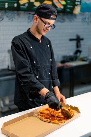 Photo for Man pizza chef cuts pizza lying in a delivery box with a pizza knife in the kitchen of pizzeria - Royalty Free Image