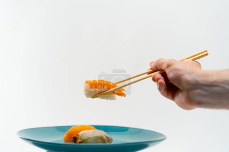 Téléchargez les photos : Gros plan d'une main masculine utilisant des baguettes pour prendre un sushi de crevettes dans une assiette de saumon et de sushi d'anguille sur fond blanc - en image libre de droit
