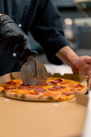 Téléchargez les photos : Homme pizza chef coupe pizza couché dans une boîte de livraison avec un couteau à pizza dans la cuisine de la pizzeria - en image libre de droit