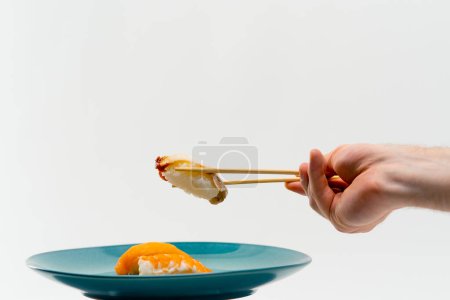 Foto de Primer plano de una mano masculina usando palillos para tomar un sushi de anguila de un plato de salmón y sushi de camarón sobre fondo blanco - Imagen libre de derechos