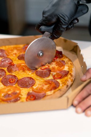 Téléchargez les photos : Homme pizza chef coupe pizza couché dans une boîte de livraison avec un couteau à pizza dans la cuisine de la pizzeria - en image libre de droit