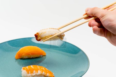 Téléchargez les photos : Gros plan d'une main masculine utilisant des baguettes pour prendre un sushi d'anguille dans une assiette de sushi de saumon et de crevettes sur fond blanc - en image libre de droit